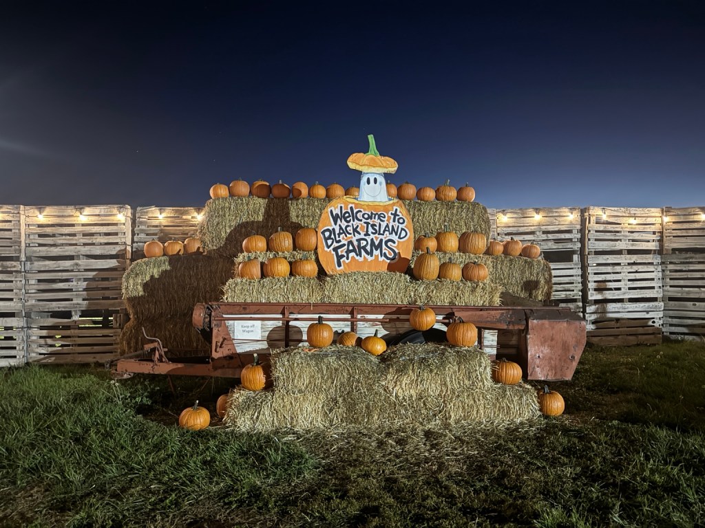 Black Island Farms welcome sign surrounded by pumpkins and hay bales on a wagon at night