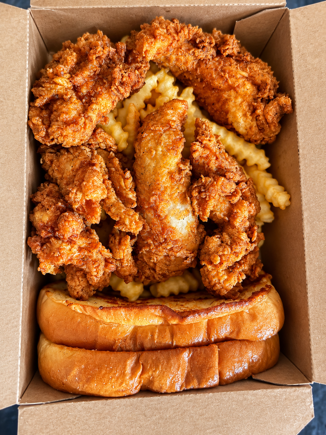 Golden fried chicken fingers with crinkle-cut fries and toast in an open takeout box