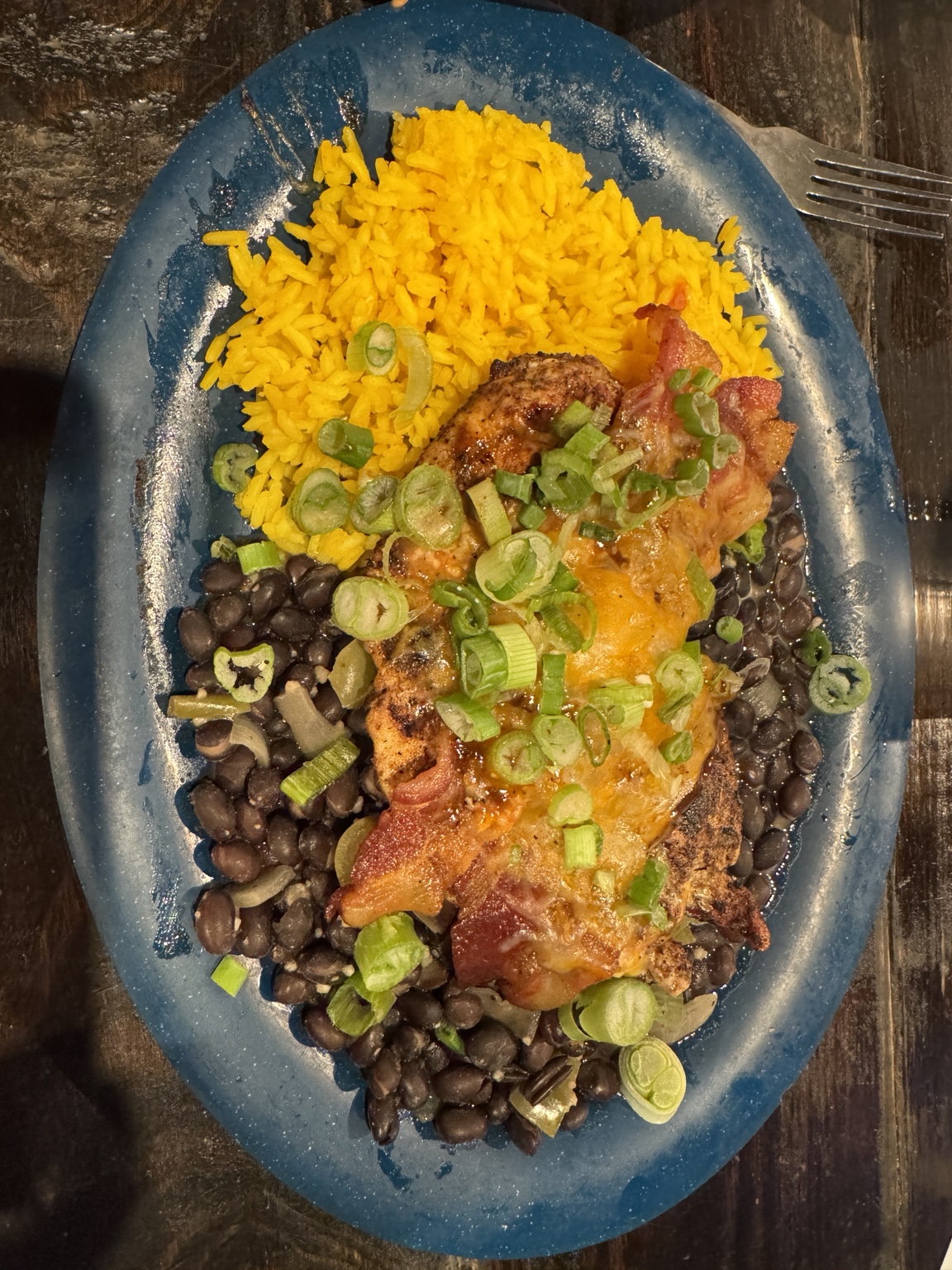 Grilled chicken topped with melted cheese, bacon, and green onions, alongside yellow rice and black beans